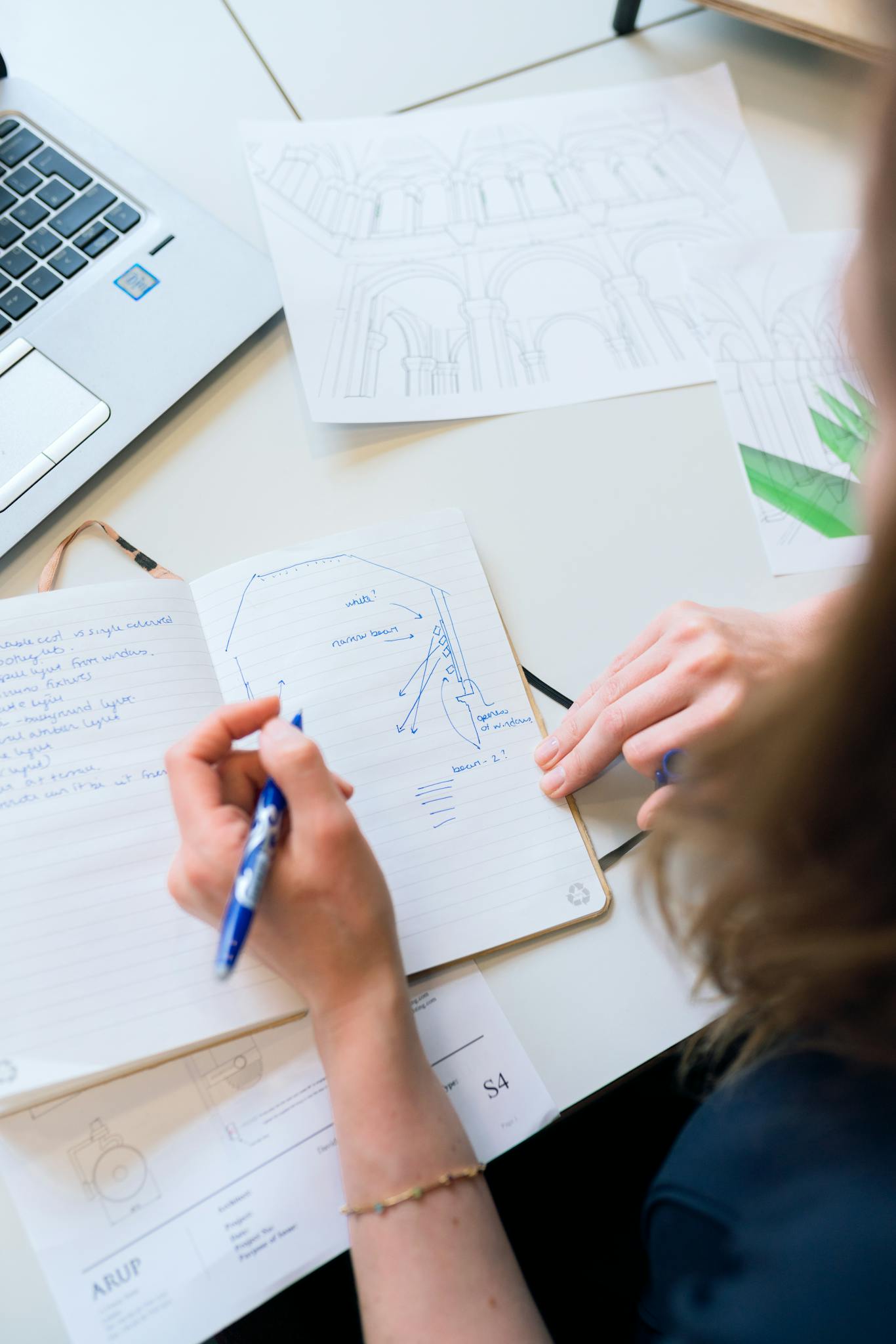 Female designer sketching architectural plans with notes and laptop in a creative workspace.